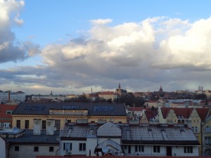 Imagem da cidade de Praga tirada do alto, aparece o telhado das casas. O céu está azul com nuvens espessas