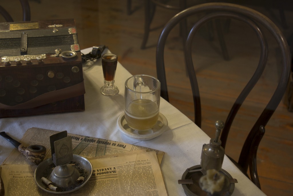 objetos de museu em cima de uma mesa: jornal, cinzeiro, copo com cerveja no final, uma dose de rum do lado