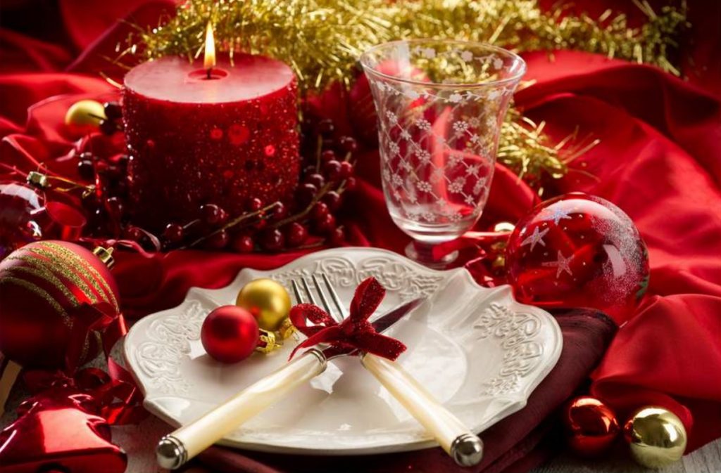 mesa de fim de ano preparada especialmente com uma toalha vermelha, vela acessa vermelha, bolas de Natal e um prato branco com talheres cruzados em cima