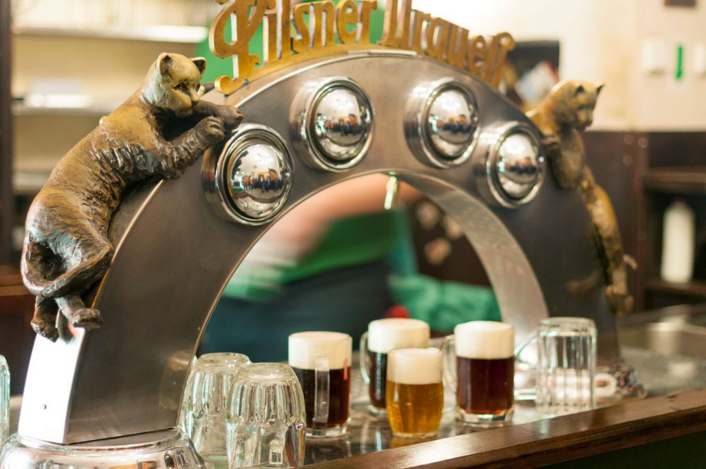 Cervejaria Duas Gatas, tradicional cervejaria localizada no centro histórico de Praga. Na foto, aparece um arco de metal com duas gatas, uma preta e outra dourada, cada uma de um lado do arco que está acoplado nas torneiras do bar para servir respectivamente uma cerveja clara e outra escura.