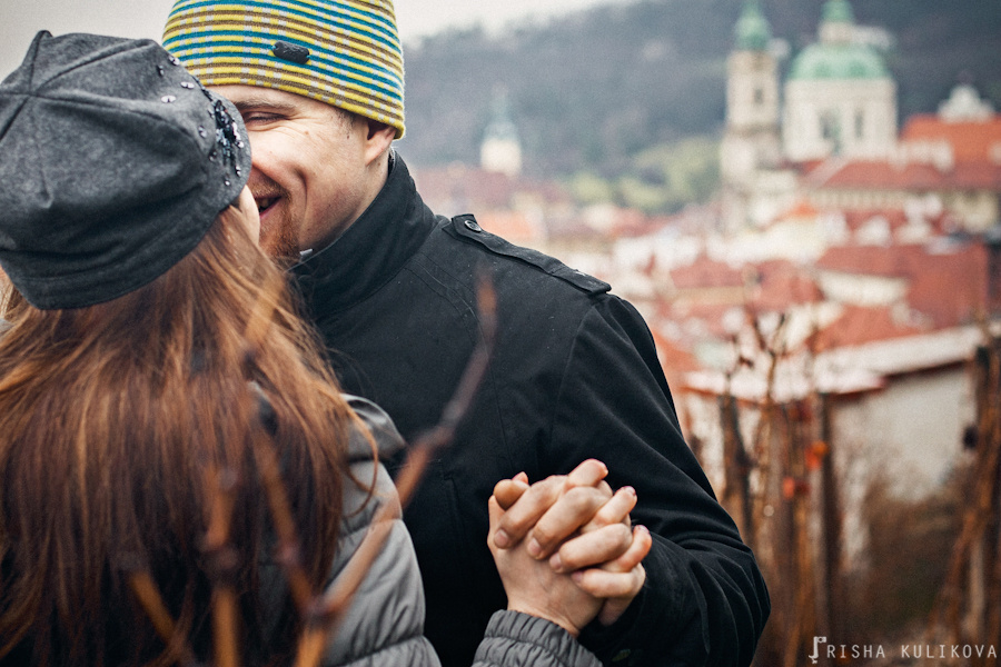Casal se beijando em close e o bairro de Malá Strana do alto ao fundo desfocado
