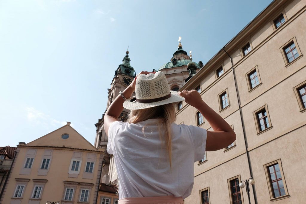 Mulher jovem com chápeu de verão, de costas para a foto, olhando para uma das torres de Praga